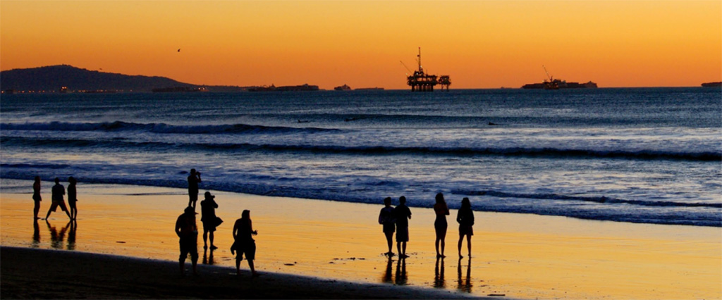 People standing on beach at sunset with oil rigs in the background